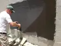 Un homme utilise une ponceuse pour araser un mur extérieur fraîchement enduit.