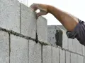 Main d'un maçon plaçant une brique sur un mur agglo, guidé par un fil à plomb.
