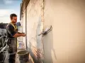 Un ouvrier applique un enduit sur agglo sur un mur extérieur, à l'aide d'une truelle. Le soleil se couche.