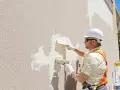 Un ouvrier en casque blanc applique un nouvel enduit sur une façade existante. Il utilise deux truelles pour refaire un enduit extérieur sur un enduit existant.