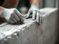 Mains gantées travaillant sur un linteau en béton brut. La texture granuleuse du matériau est visible, suggérant un travail de finition minutieux.