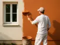 Un homme peint un mur extérieur d'une maison en orange vif avec un rouleau. Le crépi au rouleau donne une texture intéressante.