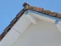 Le coin d'une toiture en pente avec des tuiles anciennes et un acrotère blanc.