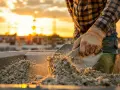Main d'un ouvrier en plein travail, manipulant du sable et des graviers pour le dosage béton. Le soleil couchant illumine la scène.