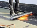 Un ouvrier réalise l'isolation d'un toit terrasse avec un chalumeau, fondant le revêtement pour une étanchéité parfaite.