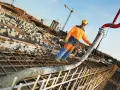 Un ouvrier en tenue orange et bleue coule du béton pour un mur banché sur un chantier. Grues et structures en construction en arrière-plan.