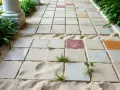 Chemin de dalles carrées sur du sable, avec des brins d'herbe poussant entre elles. L'épaisseur du lit de sable pour dalle est visible.