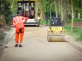 Un ouvrier prépare le chemin avec du sable stabilisé, tandis qu'un rouleau compresseur Bomag aplatit la surface.