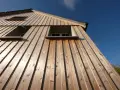 Vue en contre-plongée d'une maison avec un bardage bois clair, mettant en valeur la pose verticale des lames. Trois fenêtres sont visibles sous un ciel bleu éclatant.