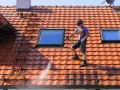 Un homme effectue le nettoyage toiture avec un nettoyeur haute pression sur un toit en tuiles rouges, sous un ciel bleu.
