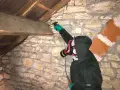Personne en combinaison et masque effectuant un traitement de charpente par pulvérisation dans un grenier en pierre.