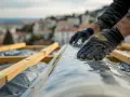 Mains gantées déroulant un pare-pluie sur une charpente en bois, avec une ville et la mer en arrière-plan.