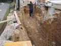 Un ouvrier travaille sur le ferraillage d'une fondation. Des armatures métalliques sont posées dans une fouille creusée dans la terre.