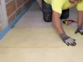 Pose d'une isolation sous dalle béton. Un ouvrier pose une feuille isolante jaune sur un sol brut, près d'un mur en briques.
