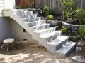 Un nouvel escalier extérieur en béton mène à un jardin paysager avec des rochers et des plantes luxuriantes.