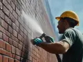 Un ouvrier en casque jaune nettoie un mur de briques avec un jet d'eau, assurant l'évacuation eau de pluie.