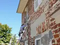 Un ouvrier sur un échafaudage réalise l'embellissement d'une façade de maison en briques rouges, sous un ciel bleu.