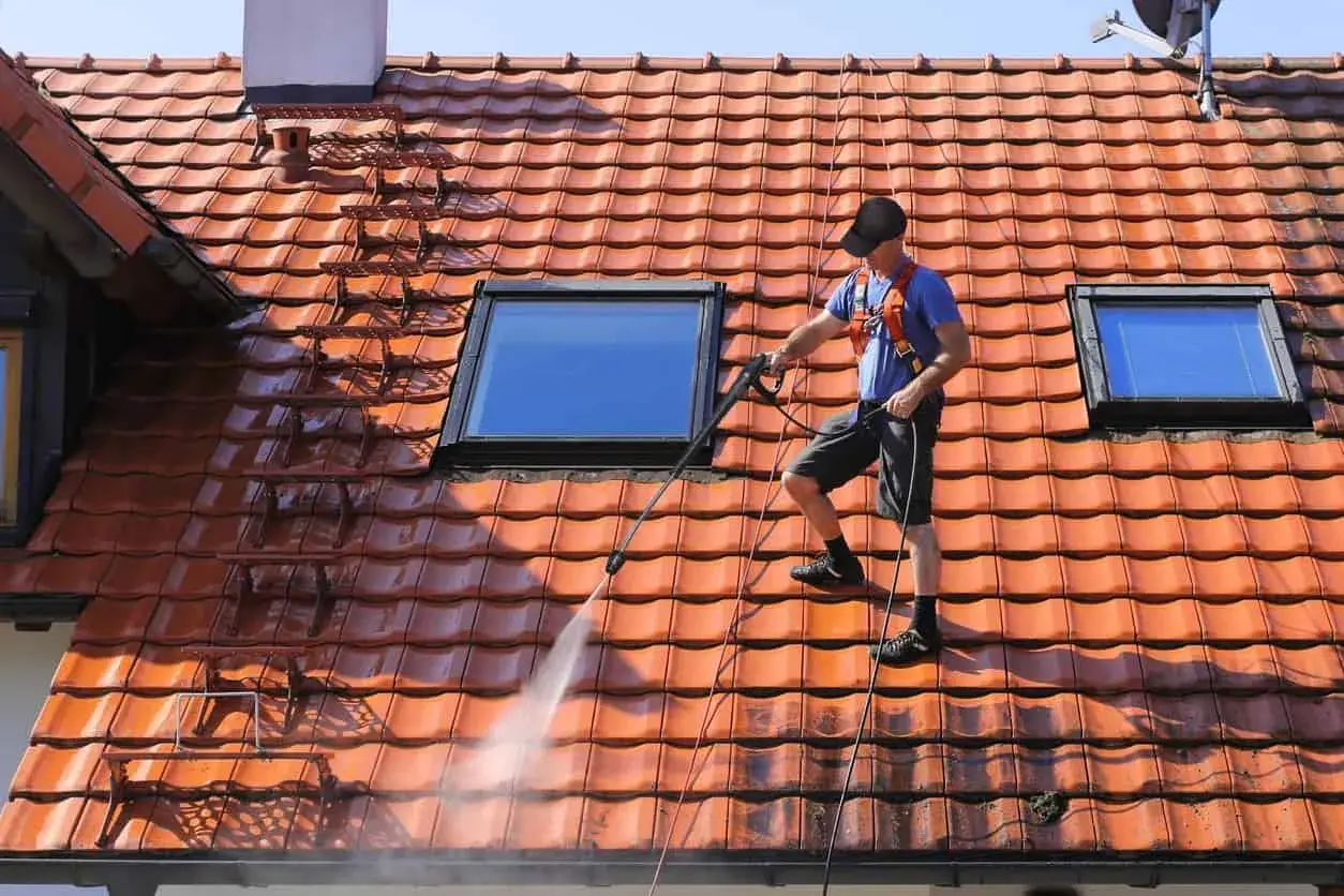 Un homme effectue le nettoyage toiture avec un nettoyeur haute pression sur un toit en tuiles rouges, sous un ciel bleu.