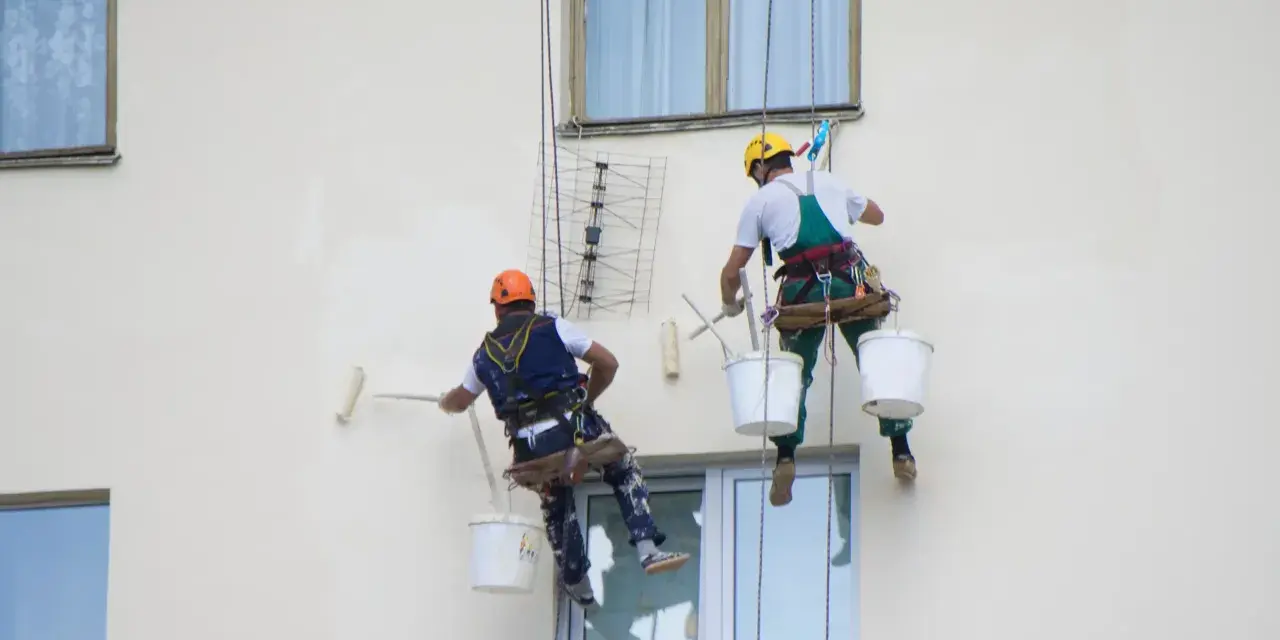 Deux peintres en bâtiment travaillent en hauteur sur une façade. L'un utilise un rouleau pour la peinture immeuble, l'autre est prêt à continuer.