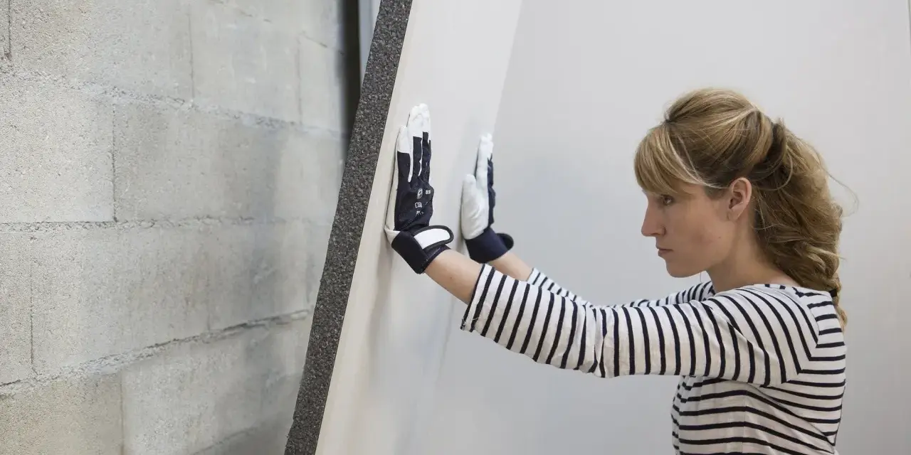 Une femme en gants manipule une plaque de doublage polystyrène expansé contre un mur de parpaings.