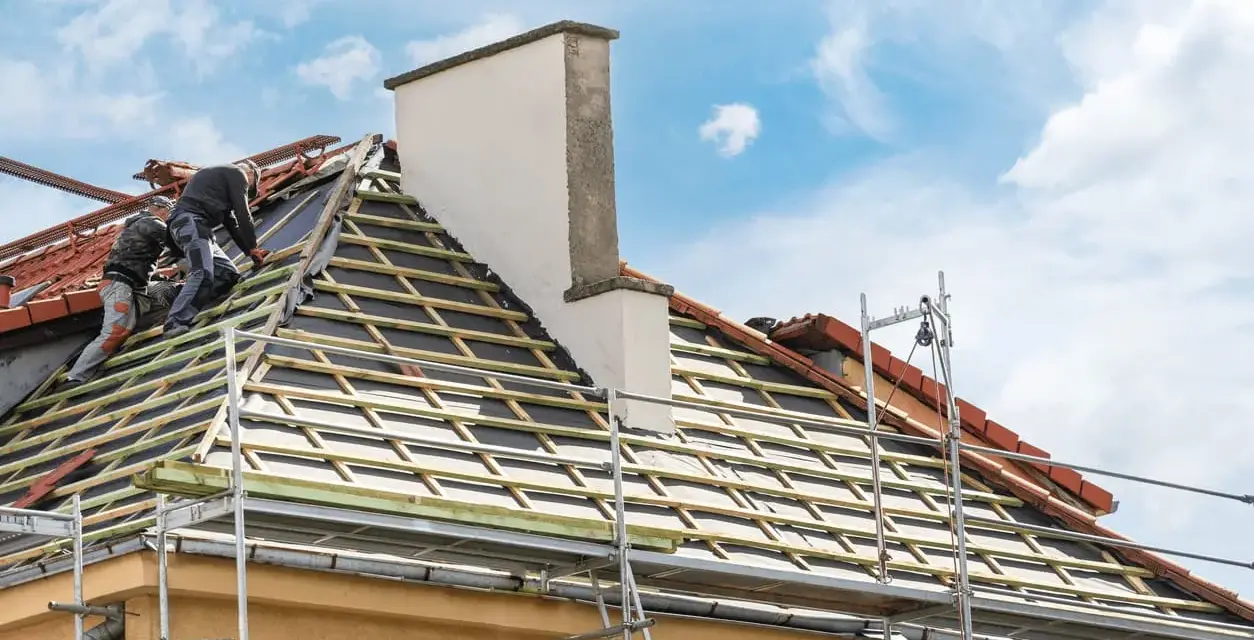 Deux couvreurs travaillent sur une toiture en cours de renovation. Des échafaudages entourent la maison.