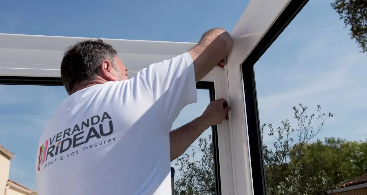 Pose d'un joint pour l'étanchéité véranda mur. Un ouvrier ajuste la structure blanche sous un ciel bleu.