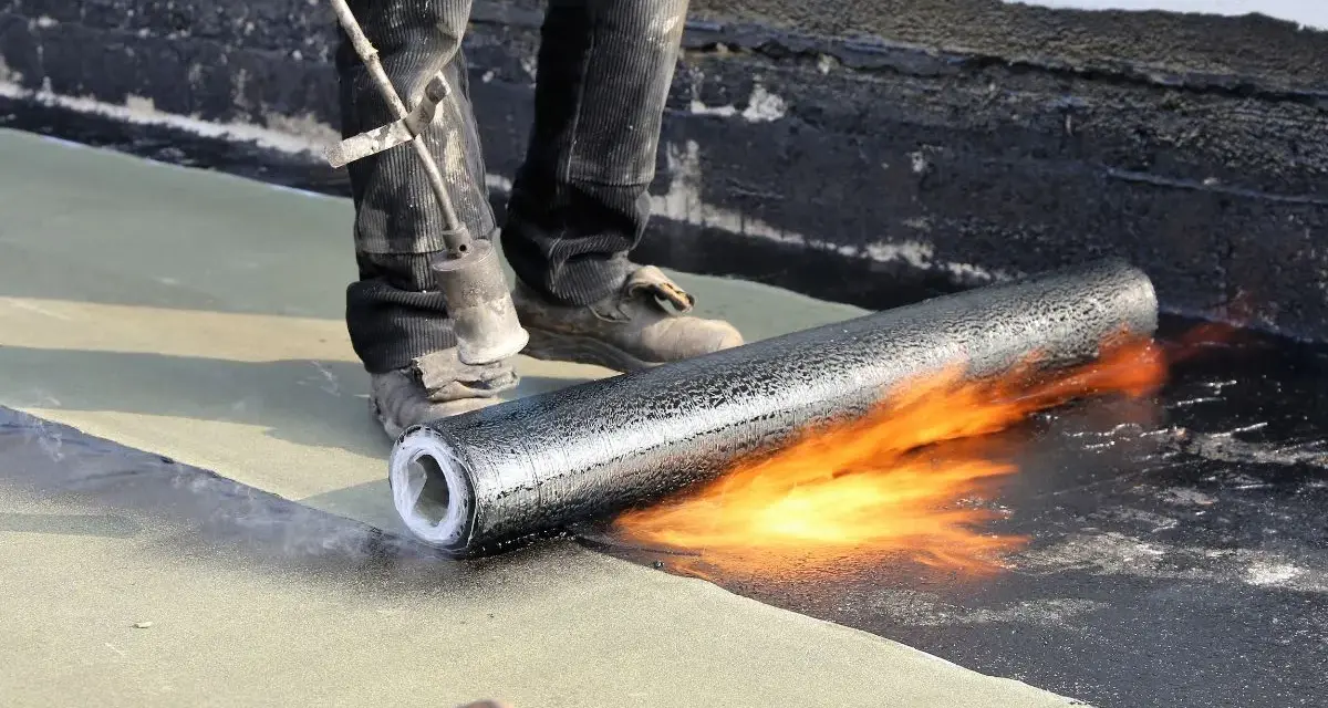 Un ouvrier réalise l'isolation d'un toit terrasse avec un chalumeau, fondant le revêtement pour une étanchéité parfaite.