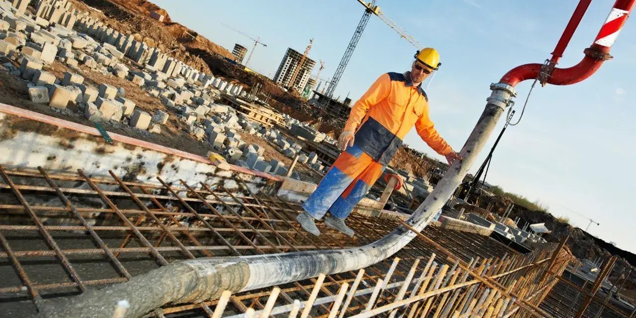 Un ouvrier en tenue orange et bleue coule du béton pour un mur banché sur un chantier. Grues et structures en construction en arrière-plan.