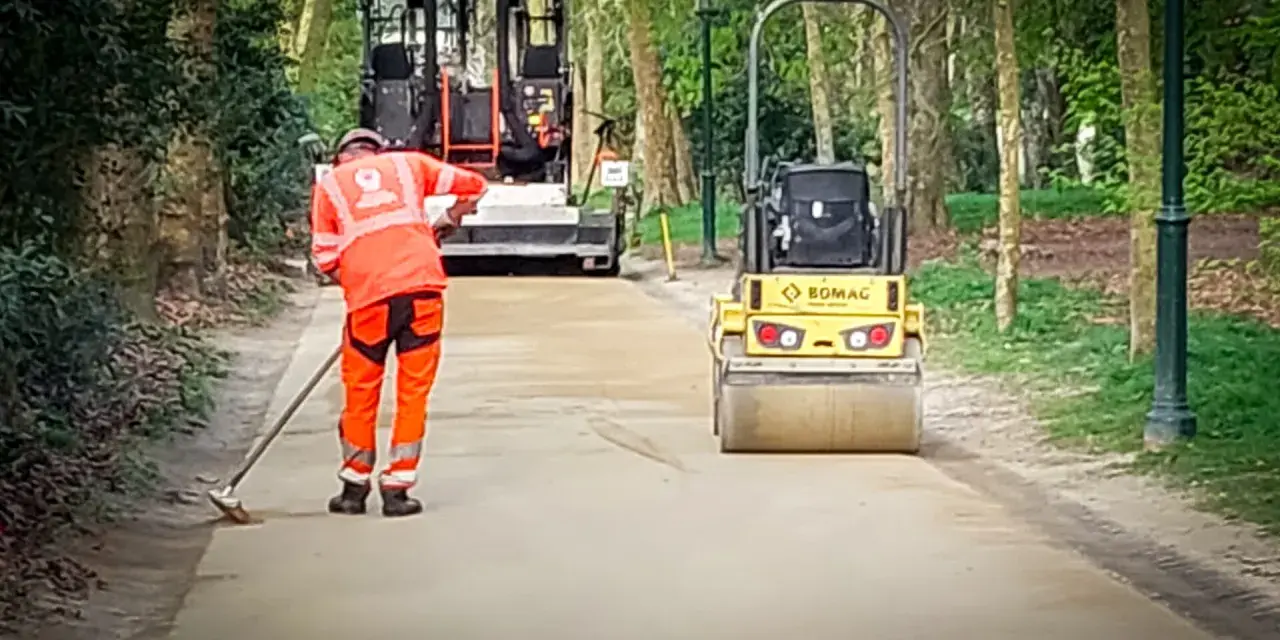 Un ouvrier prépare le chemin avec du sable stabilisé, tandis qu'un rouleau compresseur Bomag aplatit la surface.