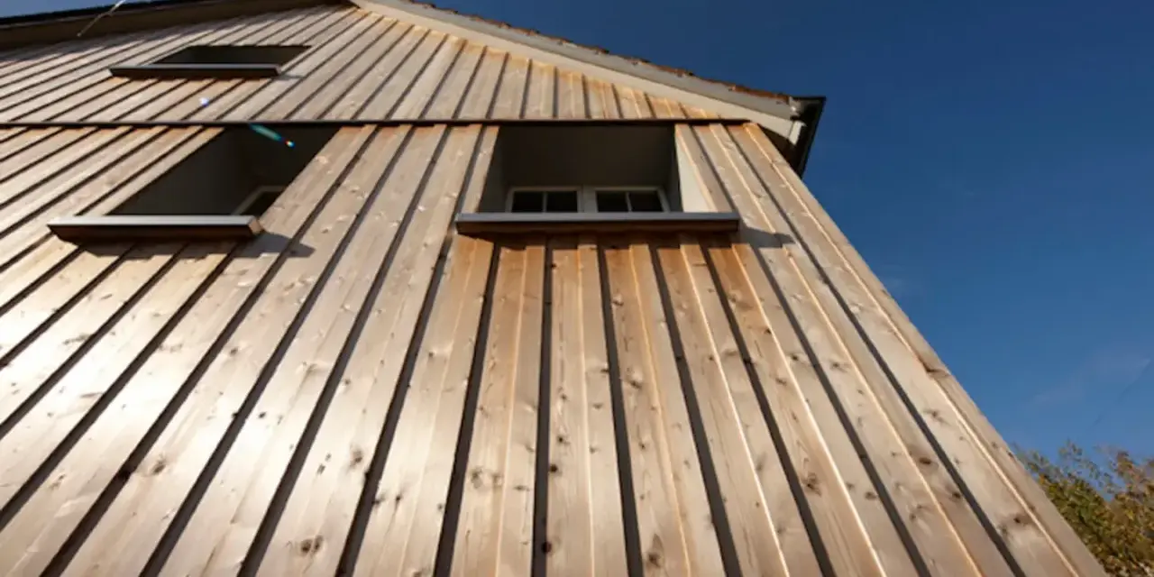 Vue en contre-plongée d'une maison avec un bardage bois clair, mettant en valeur la pose verticale des lames. Trois fenêtres sont visibles sous un ciel bleu éclatant.
