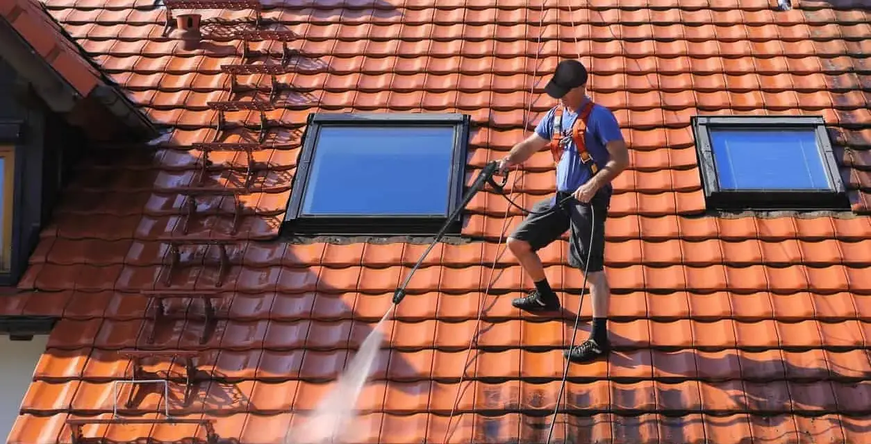 Un homme effectue le nettoyage toiture avec un nettoyeur haute pression sur un toit en tuiles rouges, sous un ciel bleu.