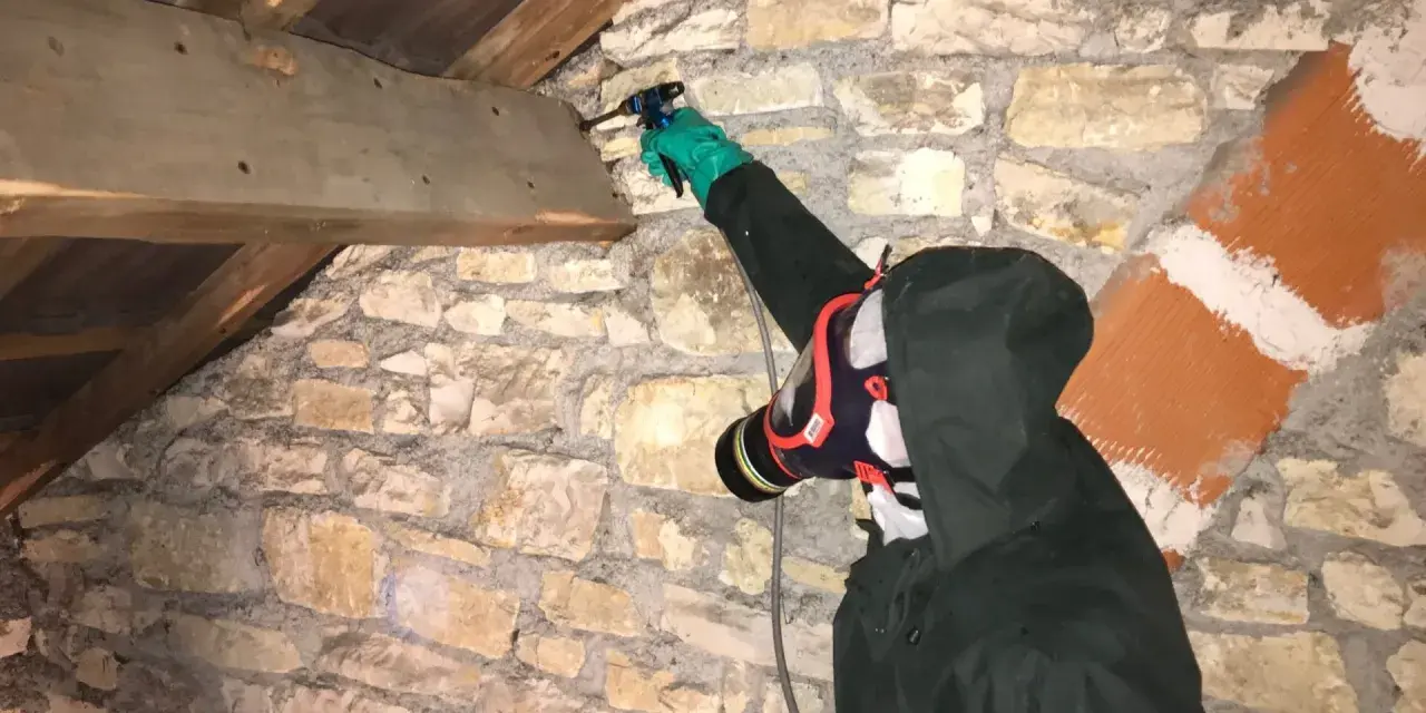 Personne en combinaison et masque effectuant un traitement de charpente par pulvérisation dans un grenier en pierre.