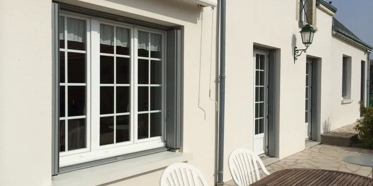 Un grand **appui de fenêtre** blanc orne la façade d'une maison. Des volets gris sont partiellement ouverts.