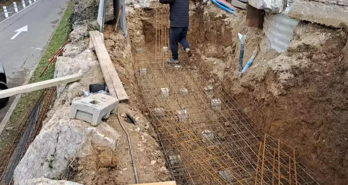 Un ouvrier travaille sur le ferraillage d'une fondation. Des armatures métalliques sont posées dans une fouille creusée dans la terre.
