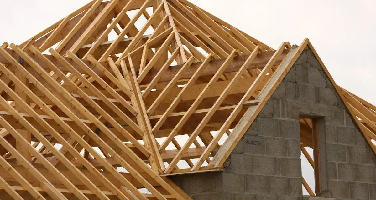 Structure en bois d'une charpente simple, en cours de construction. Les poutres de bois forment un motif complexe au-dessus des murs en blocs de béton.