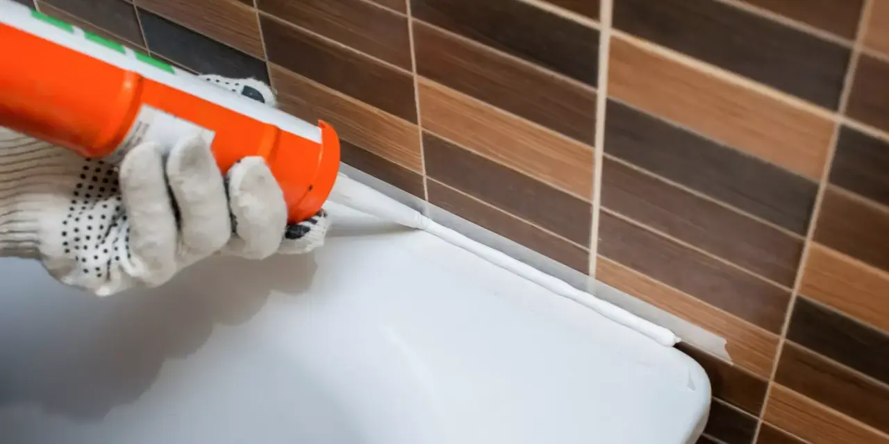 Application d'un joint acrylique ou silicone blanc pour étanchéifier le bord d'une baignoire contre un mur carrelé.