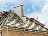 Deux couvreurs travaillent sur une toiture en cours de renovation. Des échafaudages entourent la maison.
