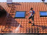Un homme effectue le nettoyage toiture avec un nettoyeur haute pression sur un toit en tuiles rouges, sous un ciel bleu.