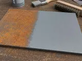 Plaque métallique rouillée d'un côté, traitée et grise de l'autre. Un pinceau et une brosse métallique montrent comment enlever la rouille.