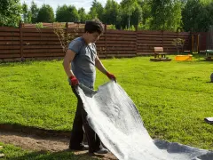 Un homme déroule un géotextile gris dans un jardin. Il réfléchit à quel geotextile choisir pour son projet d'aménagement paysager.