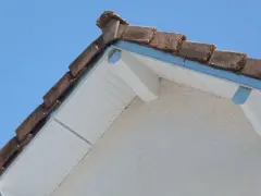 Le coin d'une toiture en pente avec des tuiles anciennes et un acrotère blanc.