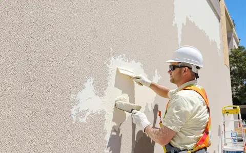 Un ouvrier en casque blanc applique un nouvel enduit sur une façade existante. Il utilise deux truelles pour refaire un enduit extérieur sur un enduit existant.