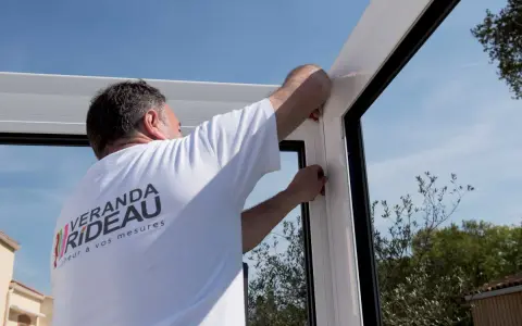 Pose d'un joint pour l'étanchéité véranda mur. Un ouvrier ajuste la structure blanche sous un ciel bleu.