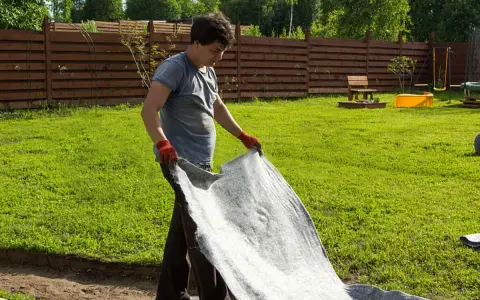 Un homme déroule un géotextile gris dans un jardin. Il réfléchit à quel geotextile choisir pour son projet d'aménagement paysager.