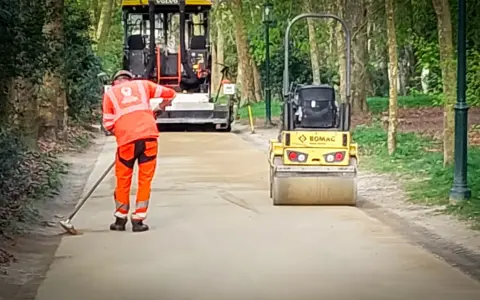 Un ouvrier prépare le chemin avec du sable stabilisé, tandis qu'un rouleau compresseur Bomag aplatit la surface.