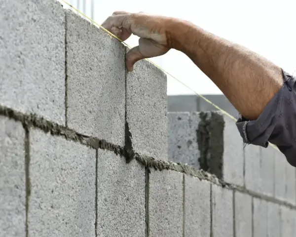 Main d'un maçon plaçant une brique sur un mur agglo, guidé par un fil à plomb.