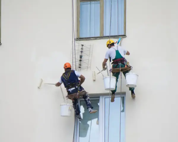 Deux peintres en bâtiment travaillent en hauteur sur une façade. L'un utilise un rouleau pour la peinture immeuble, l'autre est prêt à continuer.