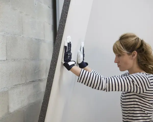 Une femme en gants manipule une plaque de doublage polystyrène expansé contre un mur de parpaings.