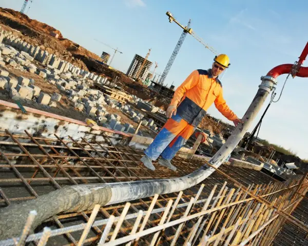 Un ouvrier en tenue orange et bleue coule du béton pour un mur banché sur un chantier. Grues et structures en construction en arrière-plan.