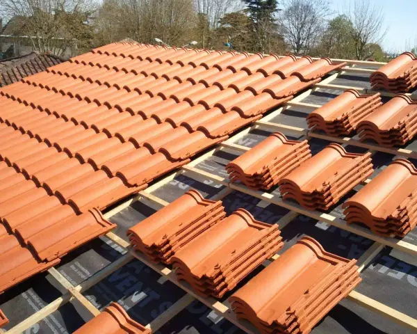 Vue d'un toit en cours de pose, révélant des rangées de tuiles mécaniques en terre cuite orange vif, empilées sur des lattes de bois.
